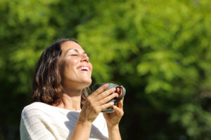 Satisfied adult woman breathing fresh air holding a coffee cup standing outdoors in the park